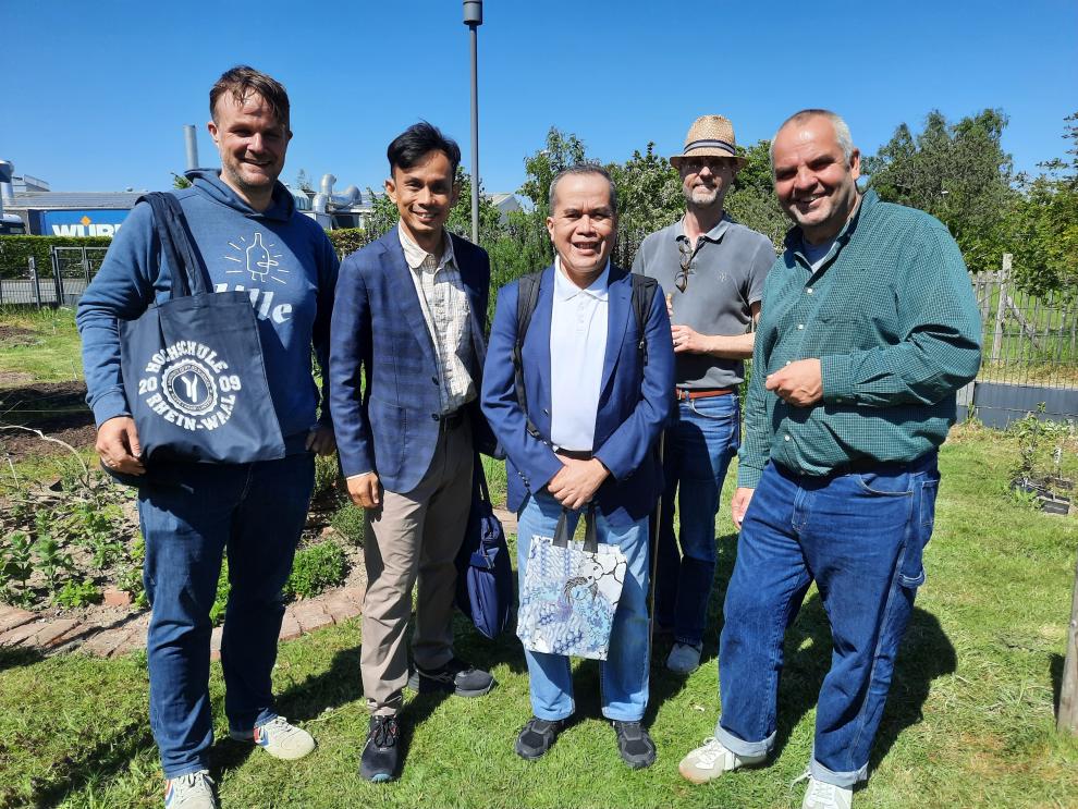 Fünf Männer stehen lächelnd nebeneinander in einem Gartenbereich des Campus. Sie tragen legere und geschäftliche Kleidung. Einige halten Stofftaschen, darunter eine mit dem Logo der Hochschule Rhein-Waal und eine weitere mit einem aufwendigen Muster. Im Hintergrund sind grüne Pflanzen und Teile eines Hochschulgebäudes zu erkennen.