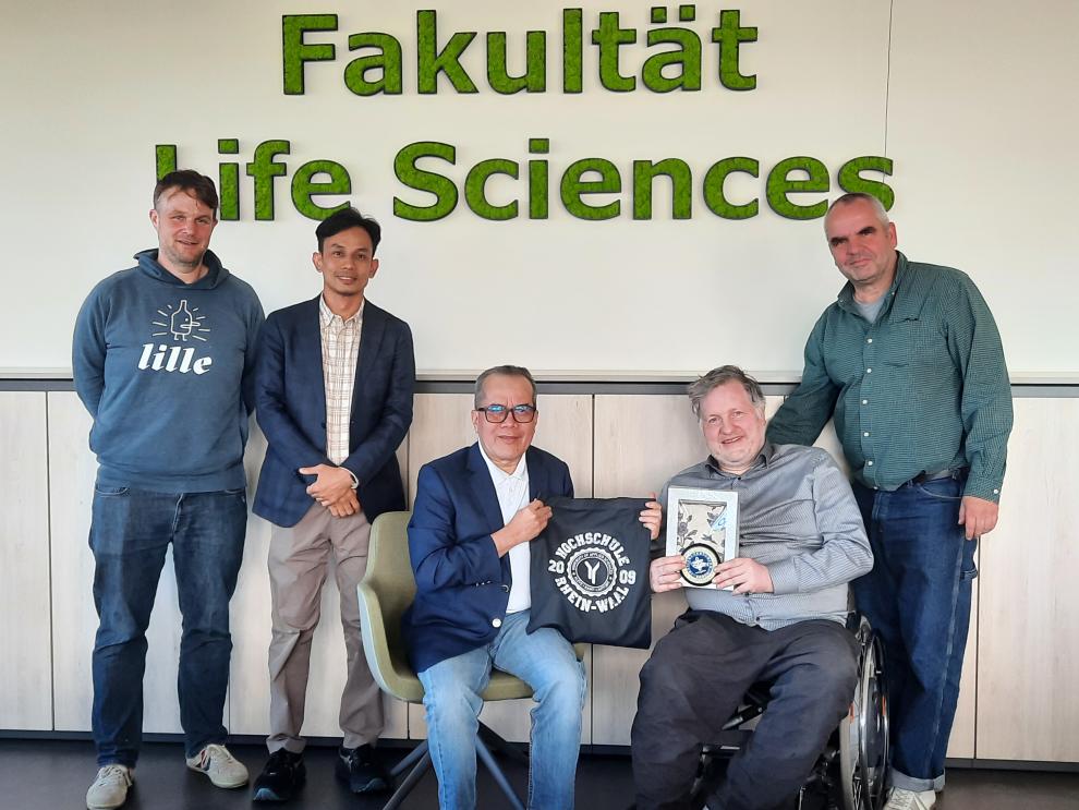 Fünf Männer posieren für ein gemeinsames Foto vor einer hellen Wand, an der der Schriftzug "Fakultät Life Sciences" aus grünem Moos angebracht ist. Einer der Männer sitzt in einem Rollstuhl. In der Mitte der Gruppe werden feierlich Gastgeschenke in die Kamera gehalten: eine blaue Stofftasche der Hochschule Rhein-Waal und ein gerahmtes Bild mit floralen Elementen.
