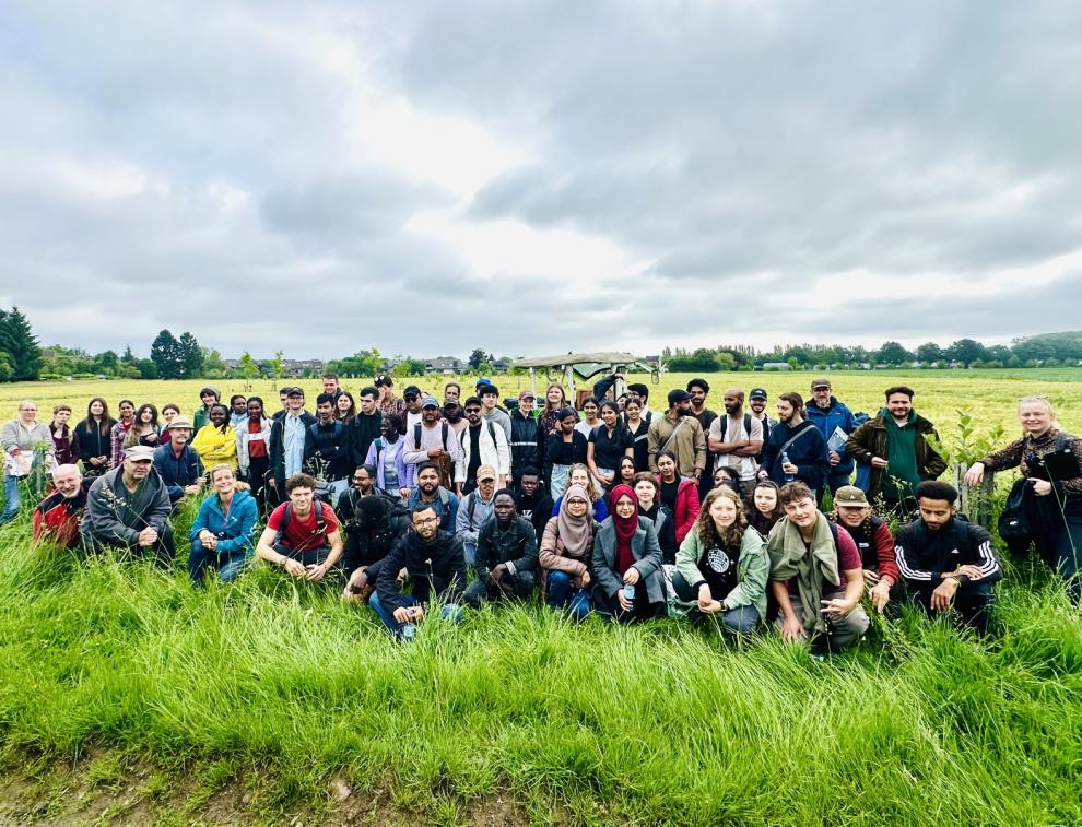 Eine große Gruppe von Studierenden und Dozierenden posiert gemeinsam in einem grünen Feld mit jungen Bäumen. Im Hintergrund steht ein grüner Traktor unter einem bewölkten Himmel.