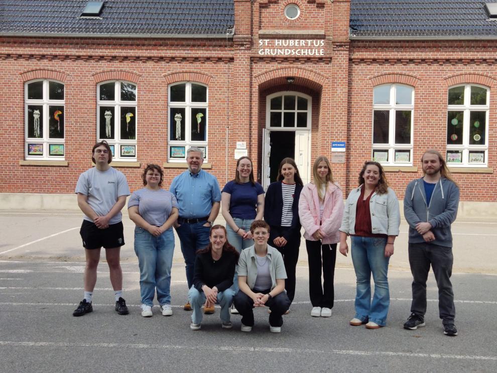 Ein Gruppenfoto von zehn lächelnden Personen – vermutlich die studentische Projektgruppe und ihr Dozent – die an einem sonnigen Tag vor dem roten Backsteingebäude der St. Hubertus Grundschule posieren.
